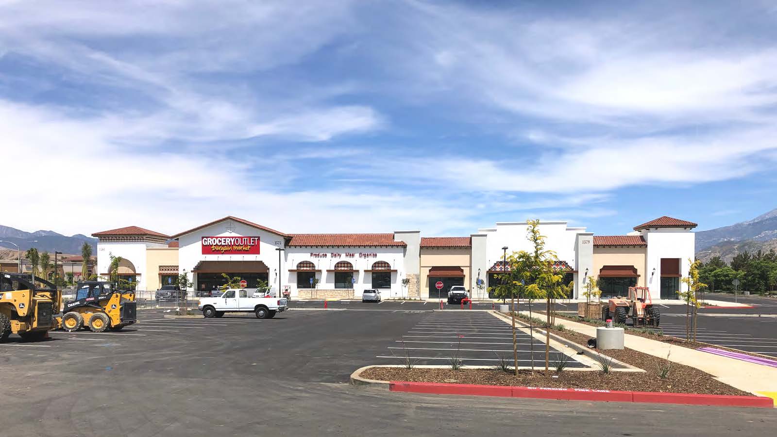 Image of Beaumont San Grogonio Village. A spacious parking lot filled with cars, with a building standing tall in the background under a clear blue sky.