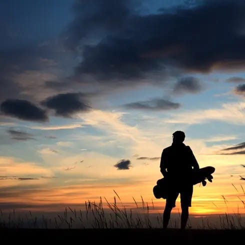 A man holding a surfboard stands in the grass, silhouetted against a vibrant sunset sky.